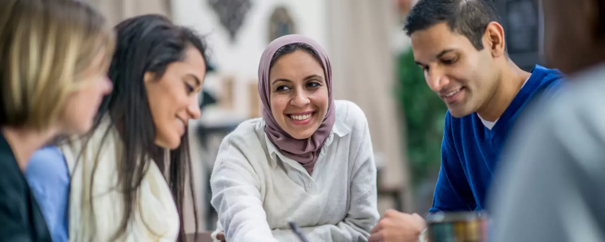 A group of people chatting at a table. Woman with hijab smiling at other women and a man in a blue sweater.