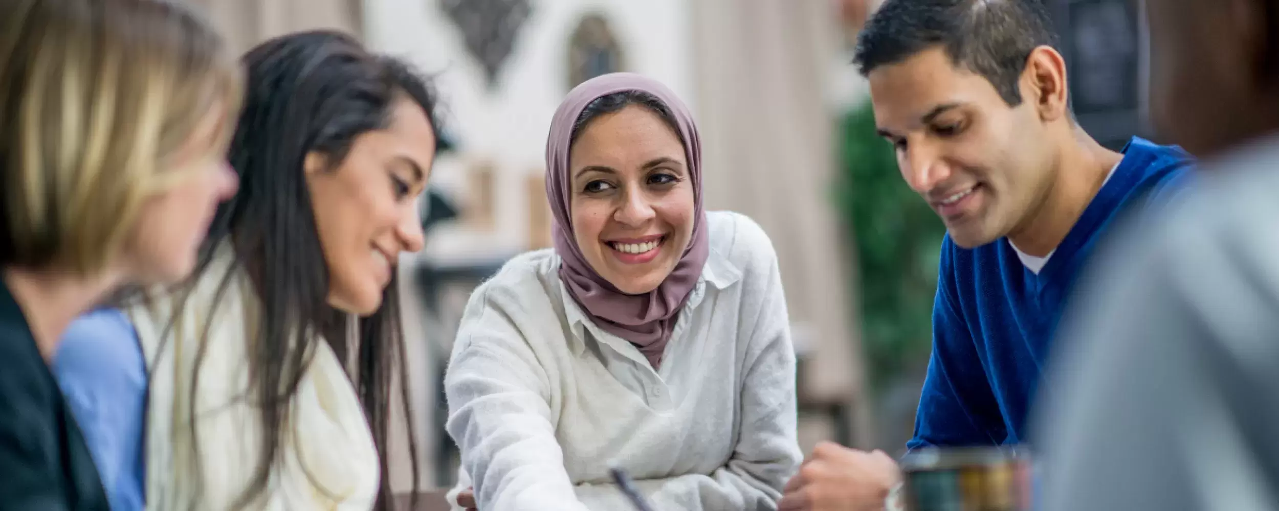 A group of people chatting at a table. Woman with hijab smiling at other women and a man in a blue sweater.